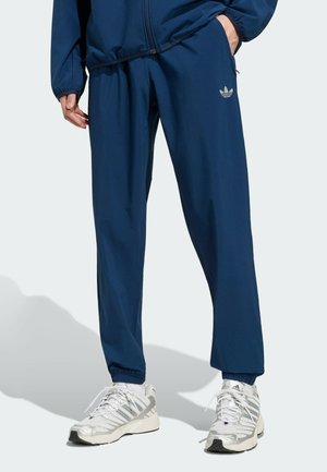 Osoba nosząca granatowe spodnie dresowe Adidas i pasującą kurtkę, zestawione z białymi i srebrnymi butami do biegania Adidas.
