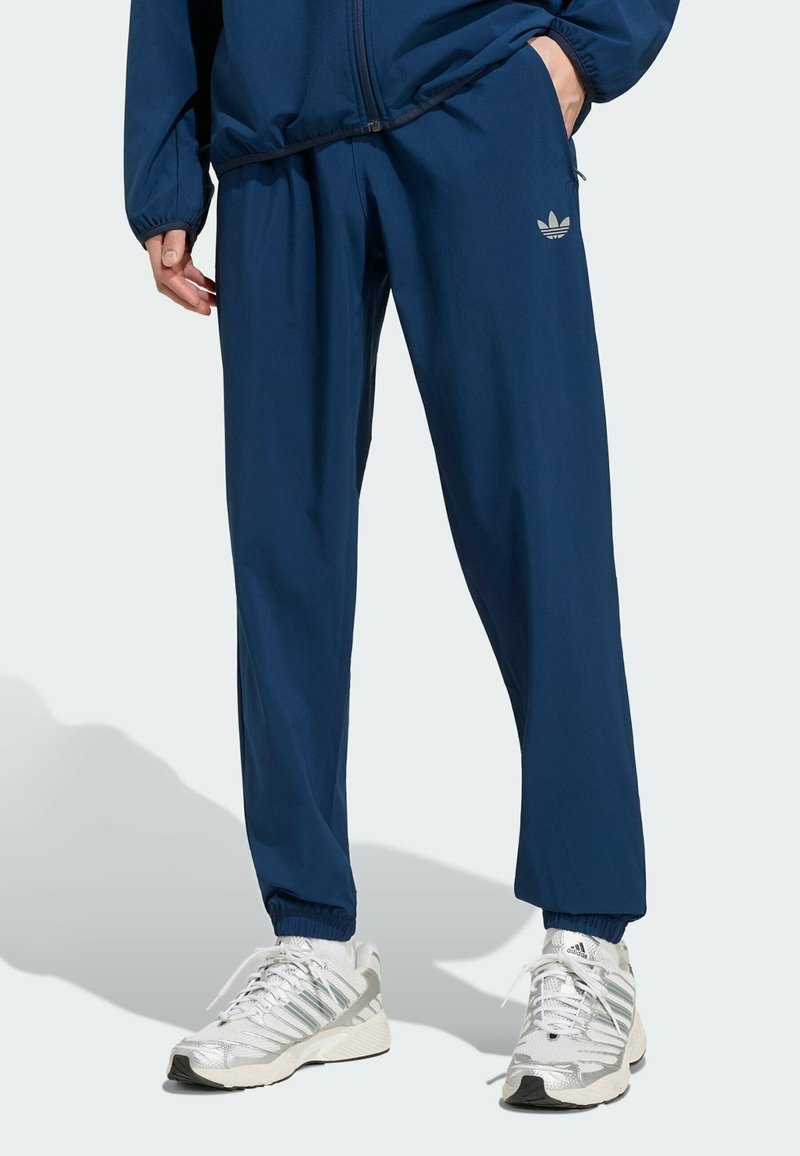 Personne portant un pantalon de survêtement Adidas bleu marine et une veste assortie, associée à des chaussures de course Adidas blanches et argentées.
