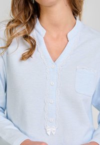 Mujer vistiendo una blusa de manga larga azul claro con borde de encaje blanco, botones blancos en el frente, un pequeño bolsillo en el pecho y un lazo blanco en la parte inferior.