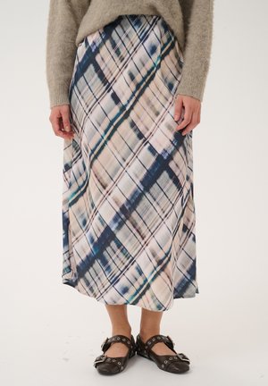 Personne portant un pull beige duveteux, une longue jupe à carreaux bleus et beiges en diagonale, et des chaussures noires à boucles, debout devant un fond uni.