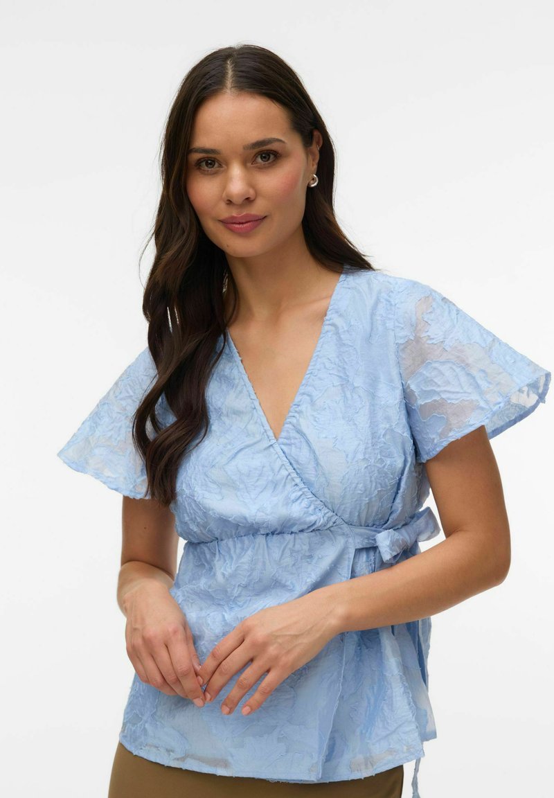 Pikkade tumejate juustega naine kannab helesinist tekstuurse mustriga ümbrispluusi lühikeste lendlevate varrukatega, seistes valge tausta ees.