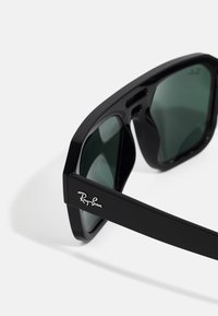 Melni plastmasas saulesbrilles ar zaļiem objektīviem. Iezīmējas ar masīvu ģeometrisku rāmi un sudraba Ray-Ban logo pie tempļa.