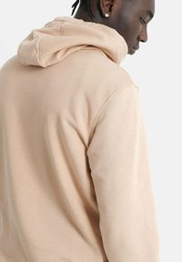 Sudadera beige con una textura suave, que presenta una capucha con cordón y un corte relajado, mostrando detalles de costura y una longitud de manga estándar.