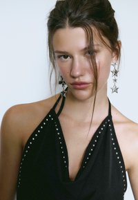 Czarny top na halter z srebrnymi ćwiekami wzdłuż dekoltu, drapowany fason, z długimi, srebrnymi kolczykami w kształcie gwiazdy. Gładka tekstura materiału.