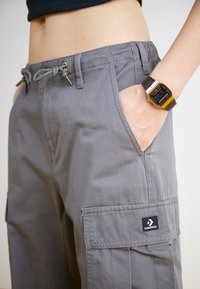 Šedé cargo kraťasy s elastickým páskem, bočními kapsami a černou nášivkou Converse. Materiál se zdá být lehký a hladký.