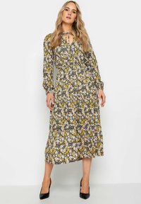 Largo vestido floral con base negra, patrones amarillos y rosas. Presenta un cuello con lazo, cintura elástica, mangas largas y falda con volantes. Tacones negros.