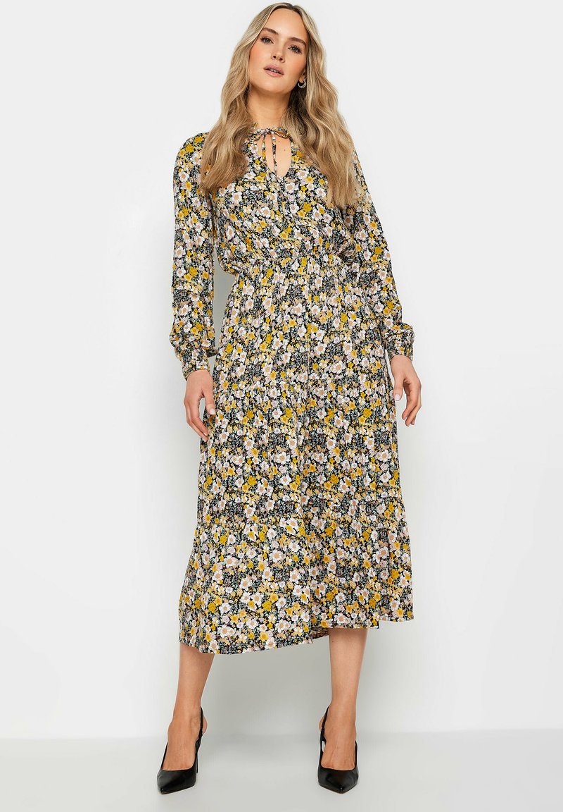 Largo vestido floral con base negra, patrones amarillos y rosas. Presenta un cuello con lazo, cintura elástica, mangas largas y falda con volantes. Tacones negros.
