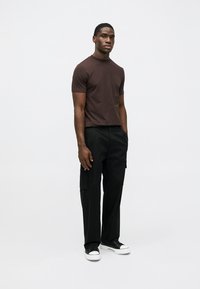 T-shirt castanha de manga curta com detalhe de bolso no peito, combinada com calças cargo pretas e ténis pretos com solas brancas.