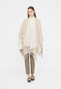Poncho beige con dettagli in frange, caratterizzato da una texture morbida. Indossato sopra un top a maglia chiaro e abbinato a pantaloni verde oliva.