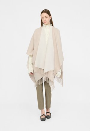 Beige poncho med fransdetaljer, med en mjuk textur. Bärs över en ljusfärgad stickad topp och matchas med olivgröna byxor.