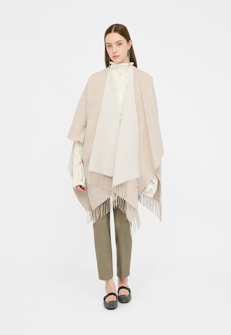 Poncho beige con dettagli in frange, caratterizzato da una texture morbida. Indossato sopra un top a maglia chiaro e abbinato a pantaloni verde oliva.