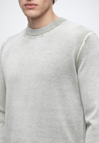 Homme portant un pull gris clair à col rond avec un petit logo brodé sur la poitrine, montré du cou jusqu'au milieu du torse.