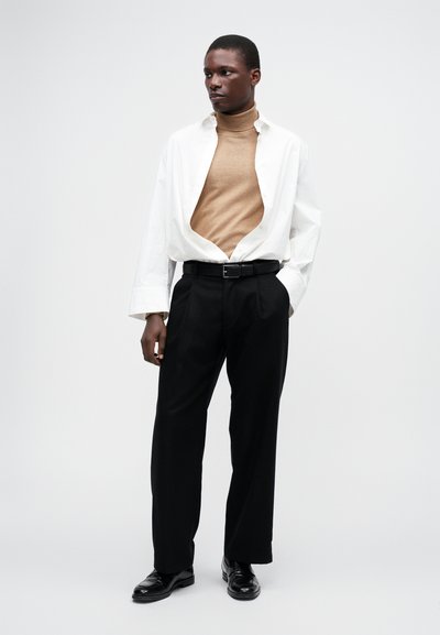 Camisa oversized branca, aberta sobre um suéter de gola alta bege, combinada com calças pretas plissadas e sapatos de couro pretos. Design simples e moderno.