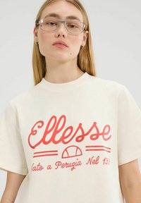 Camiseta de algodón crema con gran texto cursivo rojo "Ellesse" y un emblema gráfico. Mangas cortas y un diseño de corte relajado.