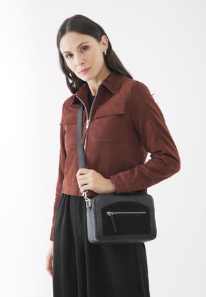 Black leather crossbody bag with a black suede front, silver zipper, and adjustable strap. Model wears a rust suede jacket and black pleated skirt.