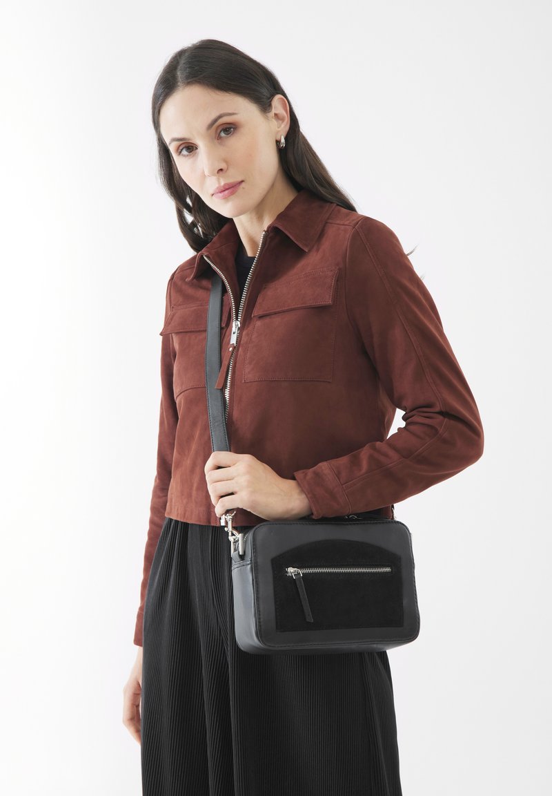 Musta nahkainen olkalaukku, jossa on musta mokka etupaneeli, hopeinen vetoketju ja säädettävä hihna. Mallilla on päällään rustinvärinen mokkatakki ja musta laskostettu hame.