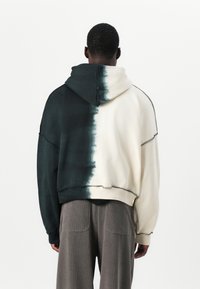 Mikina s dizajnom v color-block, kde jedna strana je tmavozelená a druhá krémová. Je vyrobená z mäkkej látky s viditeľným švom a kapucňou.