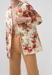 Mujer con una sudadera con cremallera floral crema sobre un suéter de tortuga beige y pantalones color camello, ajustándose la sudadera cerca del pecho.