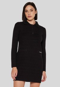 Abito nero lavorato a maglia con un motivo testurizzato, colletto abbottonato e decorazione di perline. Presenta due tasche frontali e un'etichetta con logo sul lato.
