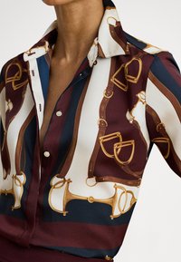 Μεταξωτό πουκάμισο με διακοσμητικό σχέδιο που περιλαμβάνει μοτίβα χαλινών αλόγου σε μπορντό, ναυτικό μπλε, κρεμ και χρυσό. Κλασικό γιακά, κλείσιμο με κουμπιά.