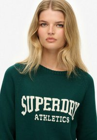 Groen gebreide trui met een brede ronde hals en de tekst "SUPERDRY ATHLETICS" in het wit over de voorkant.