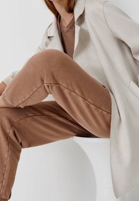 Personne assise sur un tabouret blanc portant une tenue décontractée marron et un long blazer beige clair, vue des épaules jusqu'aux jambes.