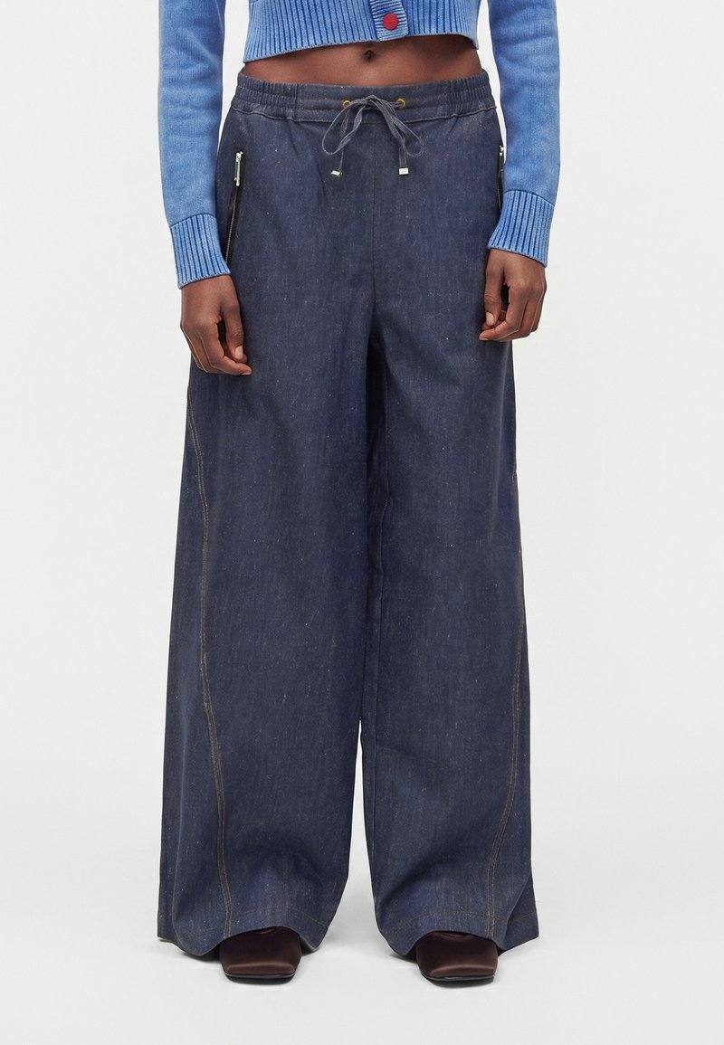 Personne portant un pantalon large sombre avec une taille à cordon, des poches zippées latérales, un pull court bleu et des chaussures sombres à bout carré.