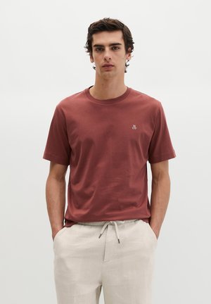 Junger Mann trägt ein rostfarbenes T-Shirt mit kleinem Logo und hellbeige Kordelzughose, steht mit den Händen in den Taschen vor einem einfarbigen Hintergrund.