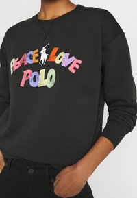 Zwarte sweatshirt met kleurrijke tekst "PEACE LOVE POLO" en een witte polo speler afbeelding. Zachte stof met geribbelde boorden en halslijn.