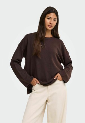 Kvinde med langt mørkt hår iført en løs mørkebrun sweater og lyse beige bukser med hænderne i lommerne mod en ensfarvet baggrund.