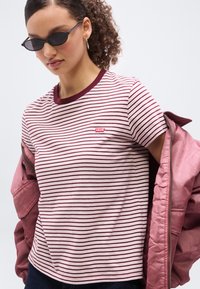 Νεαρός που φοράει σκούρα γυαλιά ηλίου, μπλουζάκι Levi's με ριγέ στο χρώμα μαρούν και λευκό, ροζ μπουφάν που γλιστράει από τους ώμους και κρίκους στα αυτιά.