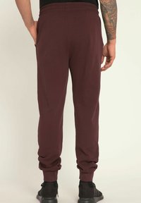 Bordowe dresy z elastycznym pasem, zwężanymi nogawkami i bocznymi kieszeniami, w zestawie z czarnymi butami sportowymi. Gładka tekstura, casualowy styl.