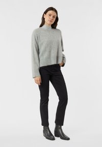 Žena stojící na bílém pozadí, oblečená v šedém svetru, černých kalhotách a černých kotníkových botách, s jednou rukou v kapse.