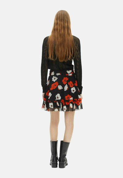 Donna con capelli lunghi, che indossa una maglietta nera a maniche lunghe con motivo, una gonna nera con fiori bianchi e rossi e stivali neri fino al polpaccio, in piedi con la schiena rivolta.