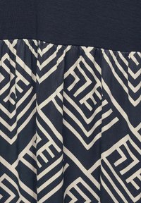 Námořnické modré šaty s žebrovanou horní částí. Sukně má bílý geometrický vzor na tmavém pozadí s nařasenou texturou.