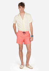 Camisa de manga corta de color amarillo claro, pantalones cortos de baño color coral con pequeños patrones blancos, complementados con unos zapatos de lona beige. Atuendo simple y casual de verano.