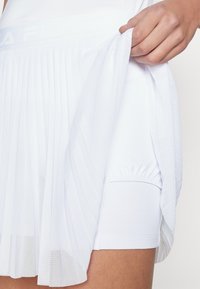White athletic skort featuring pleated design, elastic waistband, and integrated shorts. Smooth fabric with lightweight, breathable texture.