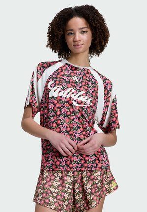 Jeune femme aux cheveux bouclés portant un t-shirt Adidas à fleurs et un short assorti à fleurs, debout avec les mains jointes à la taille.