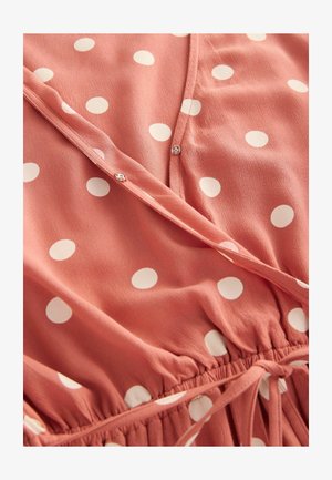 Korallfarbene Bluse mit Pünktchenmuster aus leichtem Stoff; zeichnet sich durch ein Wickeldesign, Knöpfe und eine geraffte Taille mit Bändern aus.