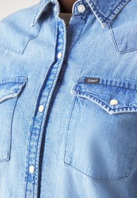 Světle modrá džínová košile s špičatým límečkem, patentkami, dvěma náprsními kapsami a malou černou visačkou "Lee" na kapse.