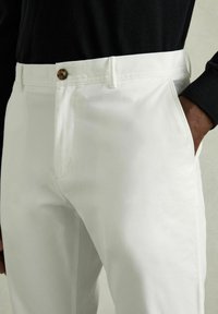 Pantaloni albi din bumbac cu front plat, buzunare laterale și închidere cu nasture. Țesătura pare netedă, cu un ușor luciu.
