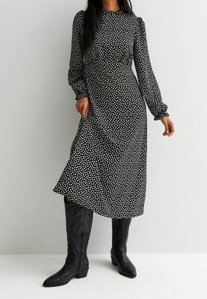 Femme portant une robe midi noire à manches longues avec des pois blancs et des bottes en cuir noires montantes jusqu'aux genoux, debout contre un mur uni.