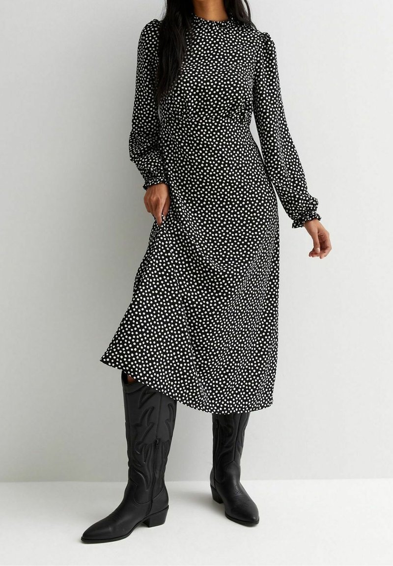 Femme portant une robe midi noire à manches longues avec des pois blancs et des bottes en cuir noires montantes jusqu'aux genoux, debout contre un mur uni.