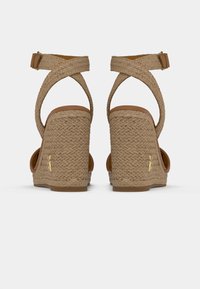 Une paire de sandales compensées tressées beiges avec des brides de cheville et des accents en logo doré sur les côtés extérieurs, vues de l'arrière.
