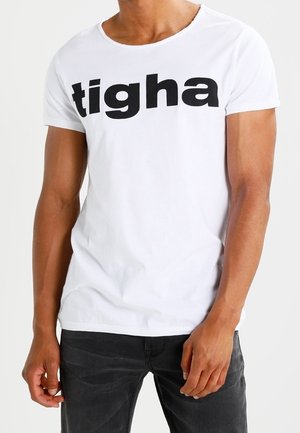 Uomo che indossa una maglietta bianca con la scritta nera in grassetto "tigha" sul petto e jeans scuri, in piedi davanti a uno sfondo semplice.