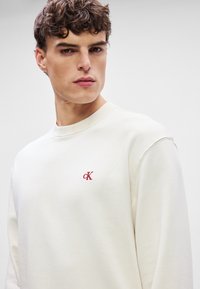 Krémová mikina s obrubou u krku, obsahující malé vyšívané logo "cK" v červené barvě na hrudi. Materiál vypadá měkce s hladkou texturou.