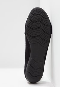 Chaussure en daim noir avec une semelle en caoutchouc texturée, dotée d'un motif ondulé et d'un logo discret, présentée en angle sur un fond blanc.