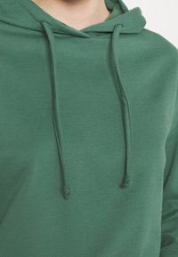 Detailní záběr osoby v zelené mikině s kapucí, na které jsou viditelné šňůrky a měkká textilní struktura.