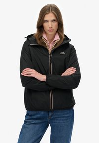 Zwarte jas met ritssluiting en capuchon, voorzien van een contrasterende ritssluiting en logo. Draagt over een lichtroze overhemd met kraag. Soepele textuur, casual pasvorm.