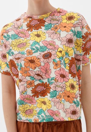 Zbliżenie na kolorową bluzkę z krótkim rękawem w kwiatowy wzór w odcieniach różu, pomarańczu, żółci i turkusu, noszoną z brązowymi spodniami.